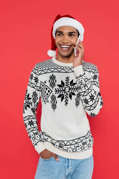Sonriente afroamericano hombre en santa hat de pie con la mano en el bolsillo mientras habla en el teléfono inteligente aislado en rojo - foto de stock