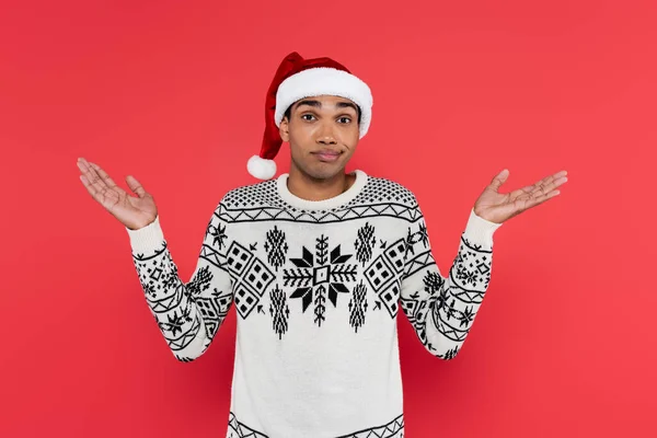 Uomo afroamericano scoraggiato in cappello di Babbo Natale e maglione bianco con motivo invernale che mostra gesto scrollata isolata sul rosso — Foto stock