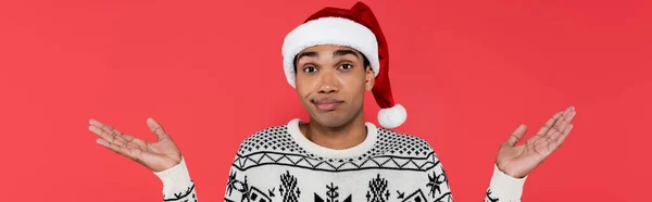 Confuso uomo afroamericano in cappello di Babbo Natale e maglione con ornamento invernale mostrando gesto scrollata isolata sul rosso, striscione — Foto stock