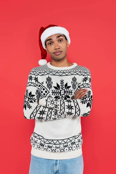 Displeased and skeptical african american man in santa hat and winter sweater standing with crossed arms isolated on red — Stock Photo