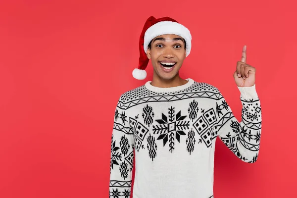 Cheerful african american man in santa hat and winter sweater showing idea gesture isolated on red — Stock Photo