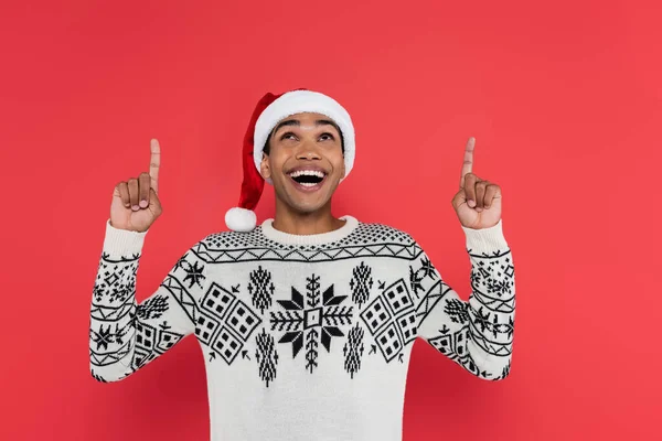 Amazed and happy african american man in santa hat and warm sweater pointing up with fingers isolated on red — Stock Photo