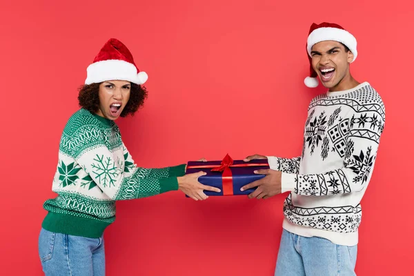 Excitada pareja afroamericana en sombreros de santa mientras sostiene la caja de regalo azul aislada en rojo — Stock Photo