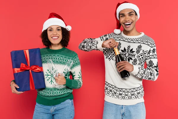 Joyful african american man opening champagne near girlfriend with blue gift box isolated on red — Stock Photo