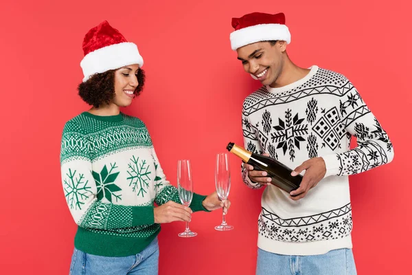 Lächelnde afrikanisch-amerikanische Frau mit Weihnachtsmütze, Gläser in der Nähe ihres Freundes, der Champagner isoliert auf Rot gießt — Stockfoto