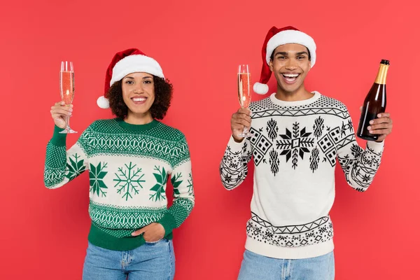 Joyful african american couple in santa hats holding champagne glasses and looking at camera isolated on red — Stock Photo