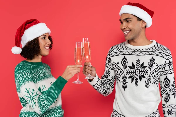 Joyful african american couple in santa hats and stylish winter sweaters clinking champagne glasses isolated on red — Stock Photo