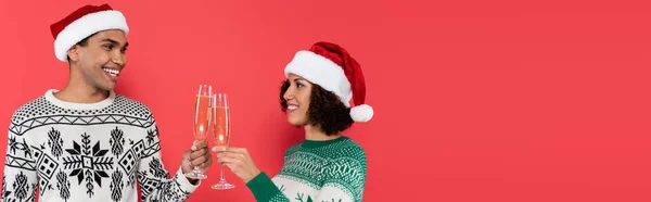 Cheerful african american couple in santa hats and warm sweaters clinking champagne glasses isolated on red, banner — Stock Photo