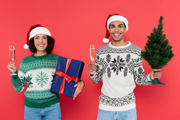 Cheerful african american couple holding champagne glasses with christmas tree and blue gift box isolated on red — Stock Photo