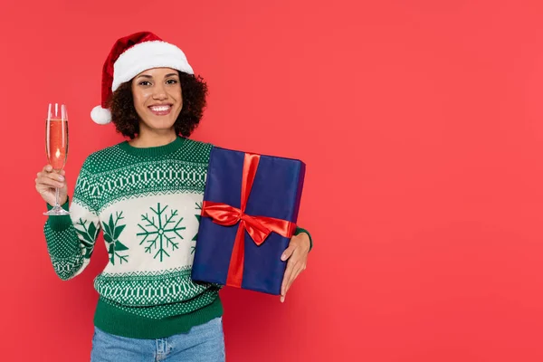 Happy african american woman in santa hat and winter sweater holding champagne glass and gift box isolated on red — Stock Photo