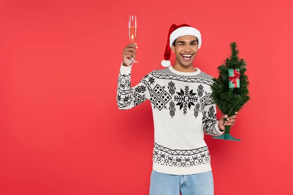 Allegro uomo afroamericano con albero di Natale e bicchiere di champagne ammiccando alla fotocamera isolata sul rosso — Foto stock