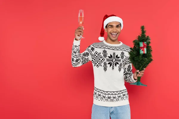 Homem americano africano alegre com árvore de natal e vidro de champanhe piscando na câmera isolada no vermelho — Fotografia de Stock