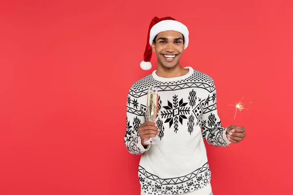 Fröhliche afrikanisch-amerikanische Mann in Weihnachtsmütze und Pullover mit Wintermuster hält Champagnerglas und Wunderkerze isoliert auf rot — Stockfoto