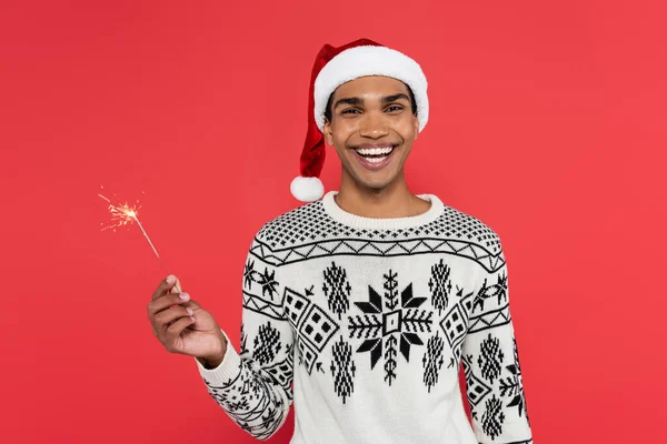 Joyful african american man in santa hat and winter sweater holding sparkler and looking at camera isolated on red — Stock Photo