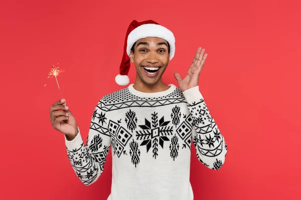 Animado afro-americano homem em santa chapéu segurando sparkler e mostrando wow gesto isolado no vermelho — Fotografia de Stock