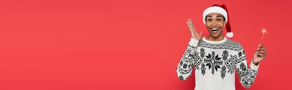 Amazed african american man in santa hat and winter sweater holding sparkler isolated on red, banner — Stock Photo