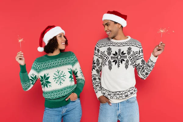 Joven afroamericano pareja en santa sombreros sosteniendo chispas y sonriendo unos a otros aislado en rojo - foto de stock