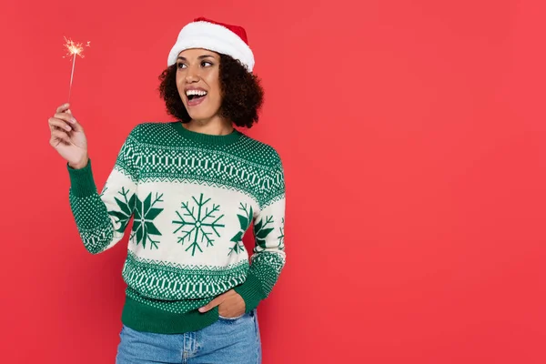 Astonished african american woman in santa hat and sweater with green pattern looking at sparkler isolated on red — Stock Photo