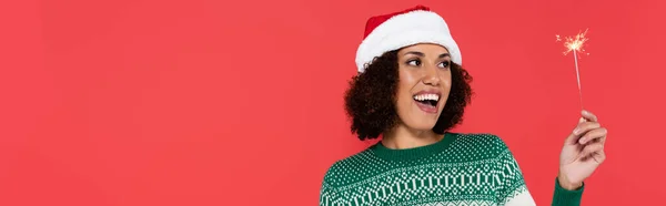 Erstaunte Afroamerikanerin mit Weihnachtsmütze blickt auf Wunderkerze auf rotem Banner — Stockfoto