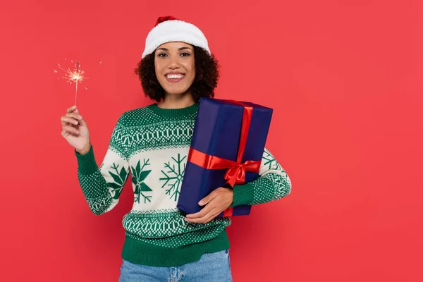 Freudige afrikanisch-amerikanische Frau in Weihnachtsmütze und Winterpullover mit Wunderkerze und blauer Geschenkschachtel isoliert auf rot — Stockfoto