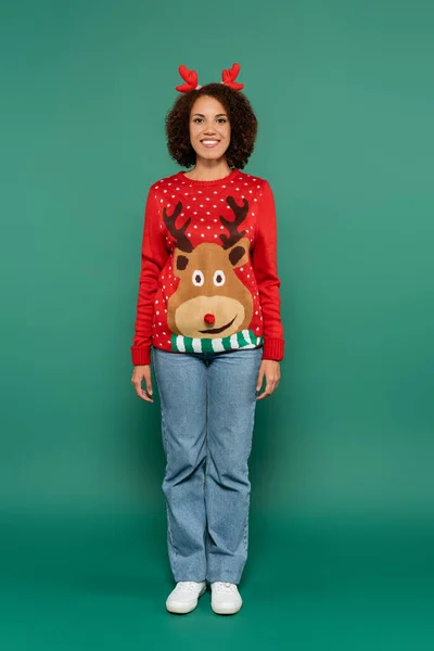 Full length of african american woman in red sweater with reindeer and in horns headband on green background — Stock Photo