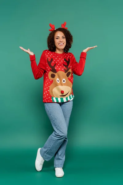 Cheerful african american woman in warm sweater with reindeer and in horns headband posing on green background — Stock Photo