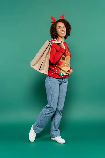 Sonriente mujer afroamericana en traje de navidad posando con bolsas de compras sobre fondo verde - foto de stock