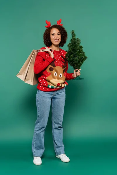 Mujer afroamericana en suéter con diadema de reno y cuernos sosteniendo árbol de navidad y bolsas de compras sobre fondo verde - foto de stock