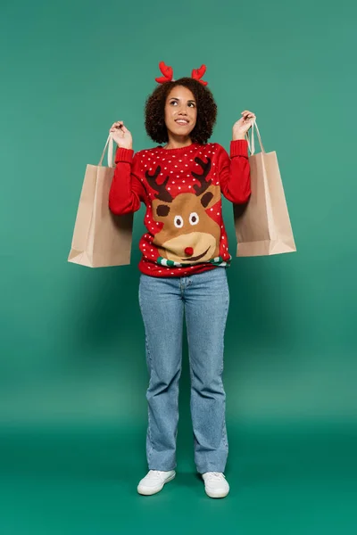 Sonriente y soñadora mujer afroamericana en traje de navidad sosteniendo bolsas de compras y mirando hacia otro lado sobre fondo verde - foto de stock