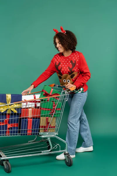 African american woman in deer horns headband taking present from shopping cart on green background — Stock Photo