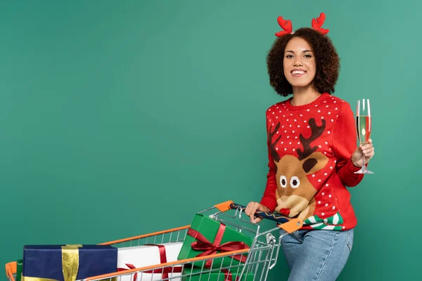 African american woman in christmas outfit holding champagne glass near shopping cart with presents isolated on green — Stock Photo