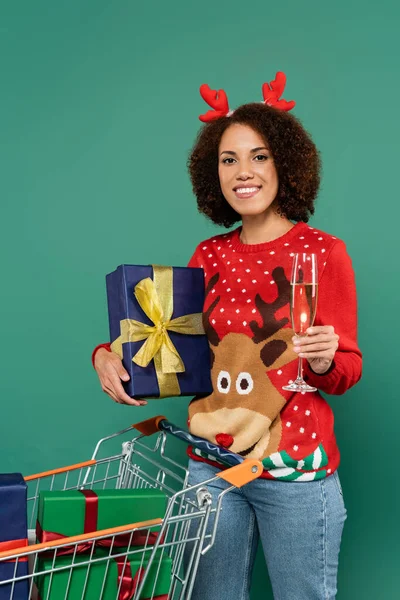 Happy african american woman in festive outfit holding champagne glass and present near shopping cart isolated on green — Stock Photo