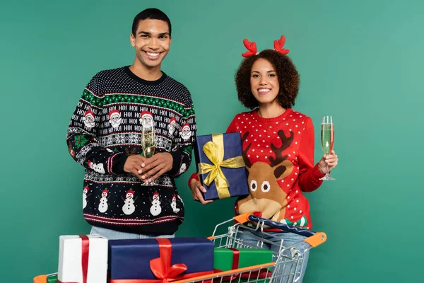 Cheerful african american couple in festive clothes standing with christmas present and champagne near shopping cart isolated on green — Stock Photo