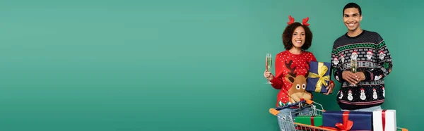 Felice coppia afro-americana in abito natalizio che tiene bicchieri di champagne vicino a scatole regalo nel carrello della spesa isolato su verde, banner — Foto stock
