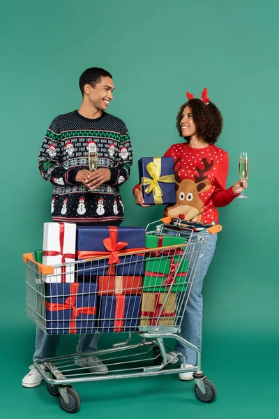 Jovem casal afro-americano com taças de champanhe sorrindo uns para os outros perto do carrinho de compras com presentes no fundo verde — Fotografia de Stock