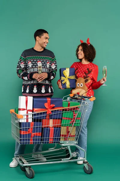 Giovane coppia afro-americana con bicchieri di champagne sorridente a vicenda vicino carrello della spesa con regali su sfondo verde — Foto stock