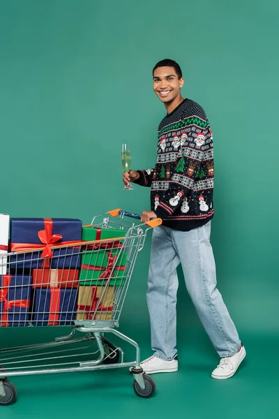 Uomo afroamericano sorridente che tiene bicchiere di champagne vicino al carrello della spesa con regali su sfondo verde — Foto stock