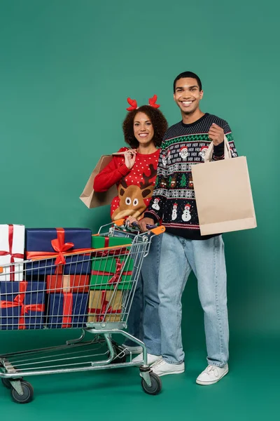 Couple afro-américain en tenue festive tenant des sacs à provisions près du chariot avec des boîtes-cadeaux sur fond vert — Photo de stock
