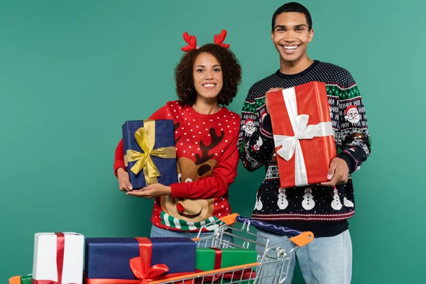 Freudiges afrikanisch-amerikanisches Paar hält Weihnachtsgeschenke in der Nähe von Einkaufswagen mit Geschenkboxen isoliert auf grün — Stockfoto