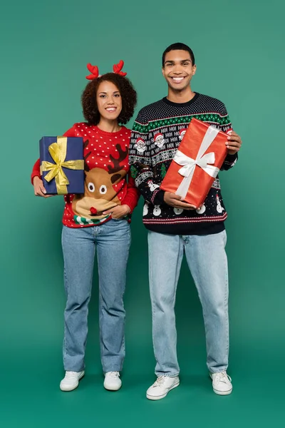 Full length of african american couple in festive outfit holding christmas presents and smiling at camera on green background — Stock Photo