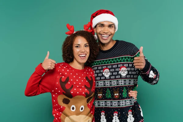 Alegre pareja afroamericana en traje de navidad mostrando pulgares hacia arriba aislado en verde - foto de stock