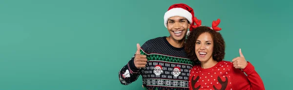 Femme afro-américaine souriante en cornes de cerf bandeau et homme au chapeau de Père Noël montrant pouces vers le haut isolé sur vert, bannière — Photo de stock