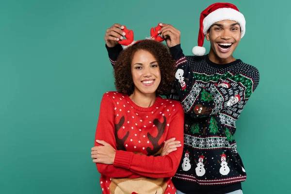 Excited african american man in santa hat touching reindeer horns on headband of girlfriend isolated on green — Stock Photo