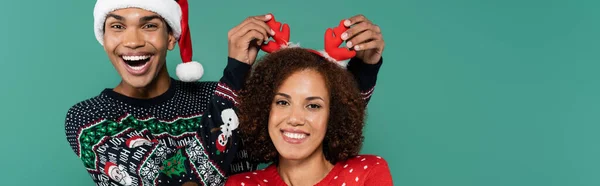 Homme afro-américain joyeux touchant cornes de cerf sur bandeau de femme souriante isolé sur vert, bannière — Photo de stock