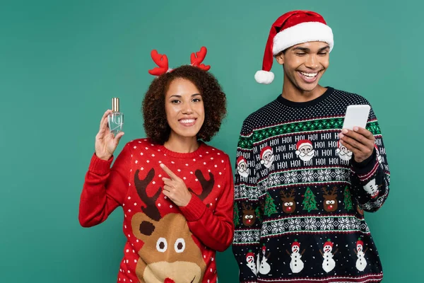 Mujer afroamericana feliz en cuernos de reno diadema apuntando a perfume cerca novio sonriente con teléfono inteligente aislado en verde - foto de stock