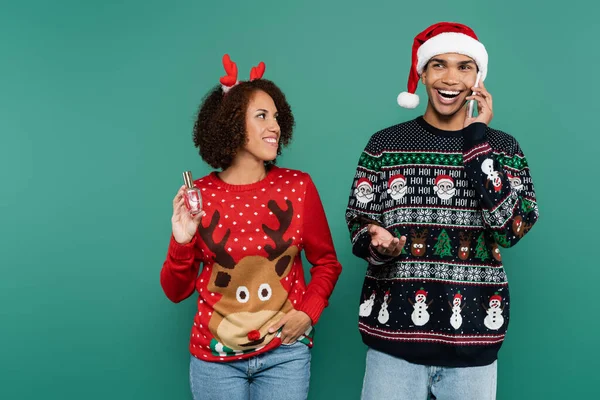 Fröhlicher afrikanisch-amerikanischer Mann mit Weihnachtsmütze, der am Handy neben lächelnder Freundin mit Parfüm auf grünem Grund spricht — Stockfoto