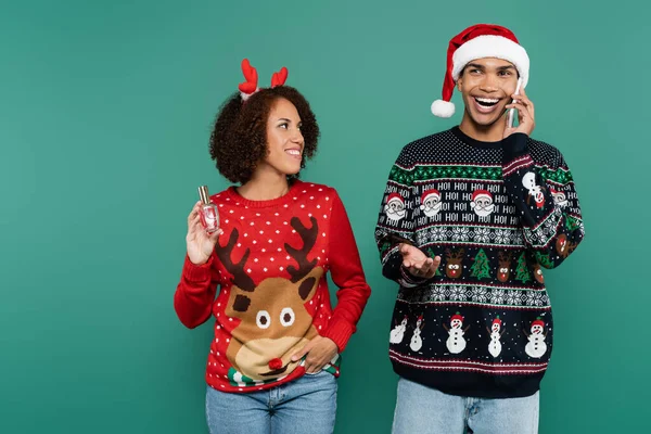 Cheerful african american man in santa hat talking on mobile phone near smiling girlfriend with perfume isolated on green — Stock Photo