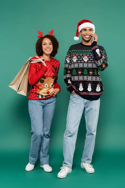 Smiling african american man in santa hat talking on smartphone near woman with shopping bags on green background — Stock Photo