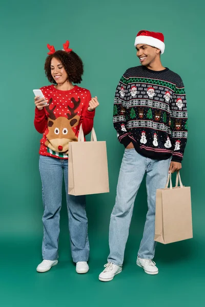Afroamerikanerin im Weihnachtskostüm blickt auf Smartphone neben lächelndem Mann mit Weihnachtsmütze auf grünem Hintergrund — Stockfoto