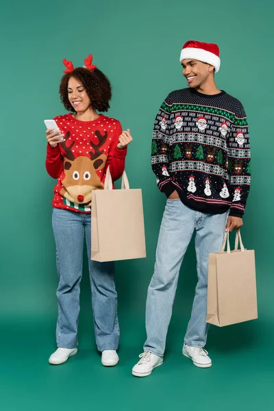 Mulher americana africana em roupa de Natal olhando para smartphone perto de sorrir homem em santa hat em fundo verde — Fotografia de Stock